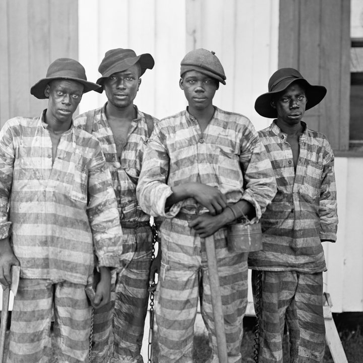 Four men in striped prison uniforms stand in front of a white wooden building.